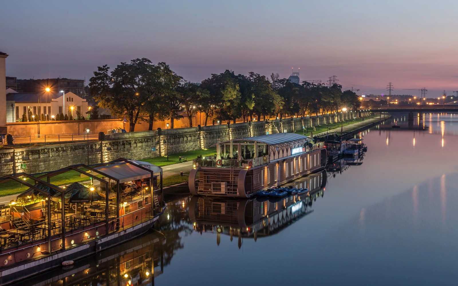 De rivieroevers van Krakau bij schemering met verlichte boten en een met bomen omzoomde promenade.