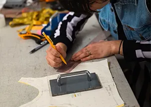 A lady is using a ruler to draw on top of a tops garment pattern on a table. there is a metal plate on top of the pattern