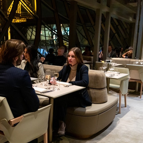 Люди сидят за столиками в современном ресторане с большими окнами и видом на освещенные структурные балки снаружи.