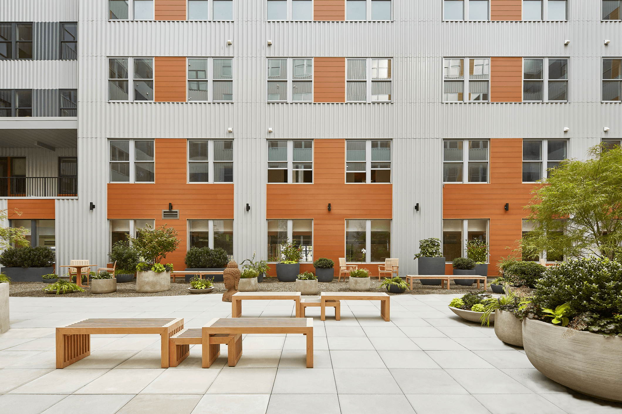 Modern building facade with orange and white accents, featuring benches and greenery in a landscaped area.