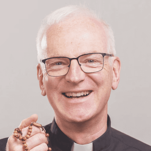 Pater Joachim Richter sthet mit einem Rosenkranz in der Hand im Priestergewand vor einer weißen Wand und lächelt
