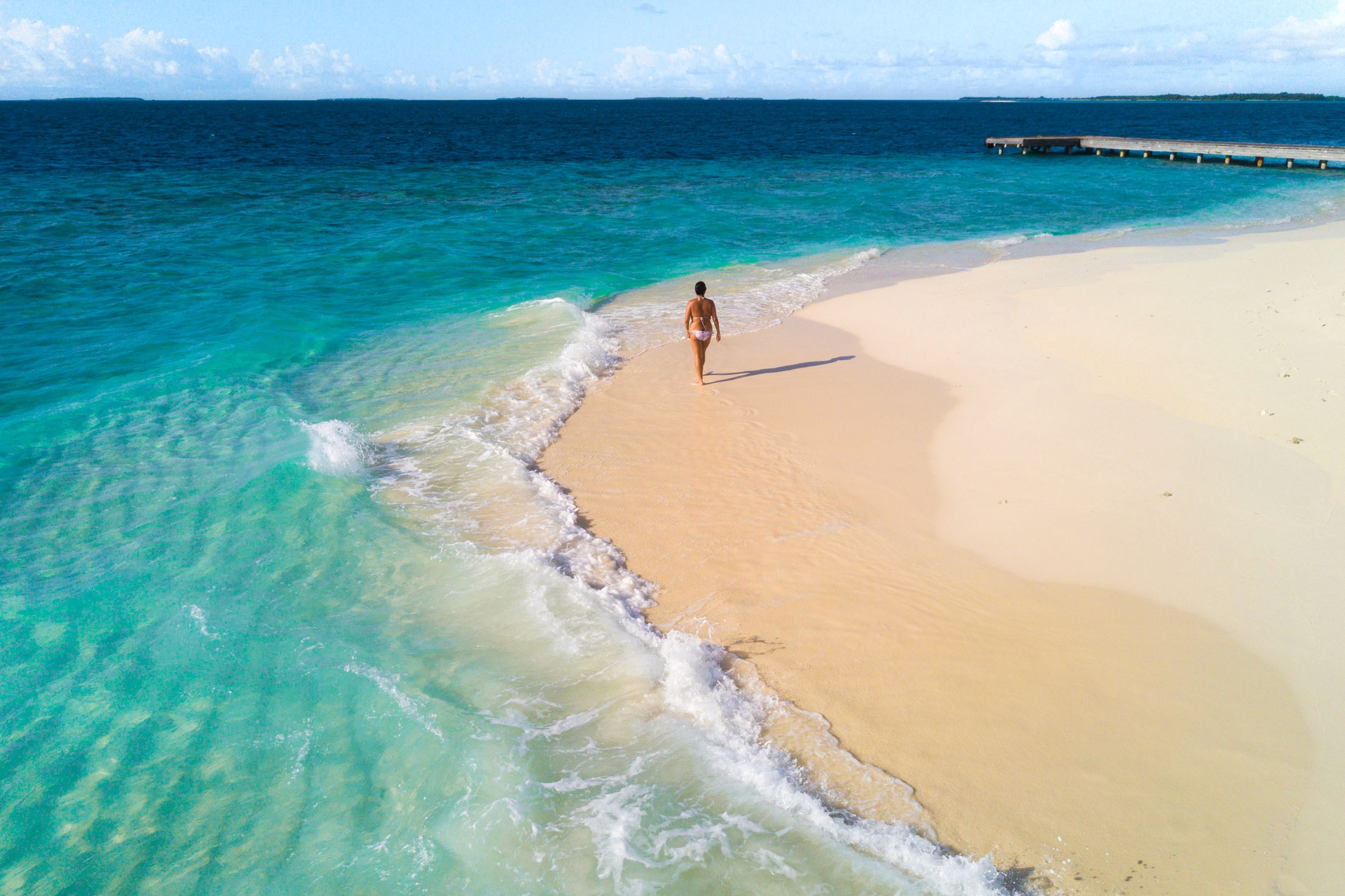 A person stands on a white sand beach next to turquoise ocean water with a pier in the distance under a blue sky.