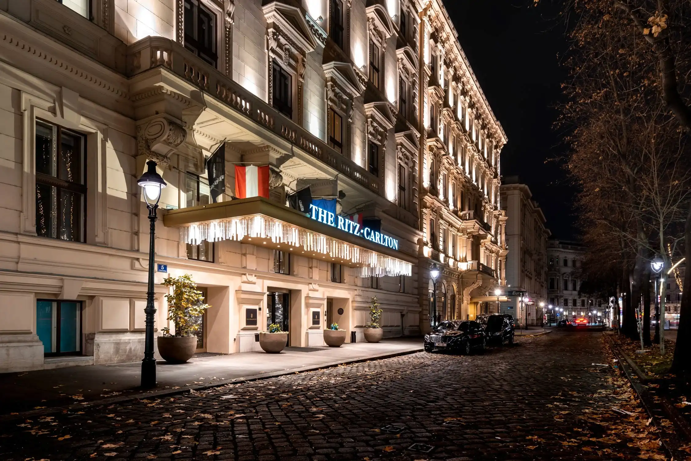 A night view of The Ritz Paris hotel with a glowing blue sign and lit windows above a cobblestone street with fallen leaves.