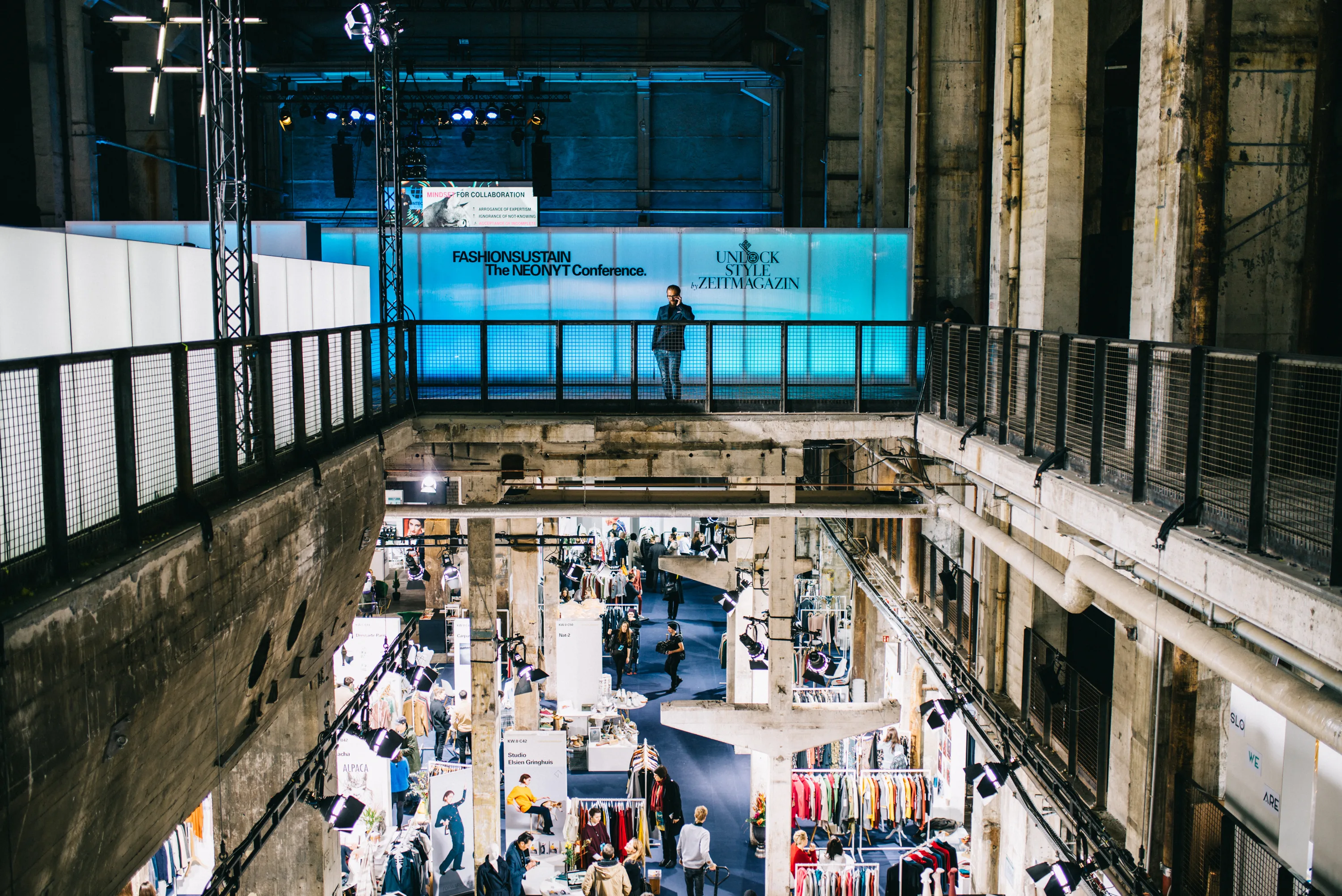 View across the Kraftwerk gallery at FASHIONTECH Berlin with an illuminated conference bridge above the exhibition floor with fashion stands.