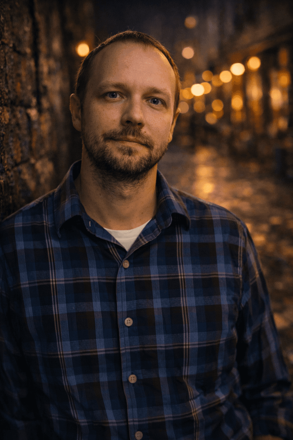 A man in a blue plaid shirt leans against a brick wall, illuminated by warm, glowing streetlights along a wet cobblestone path at night.