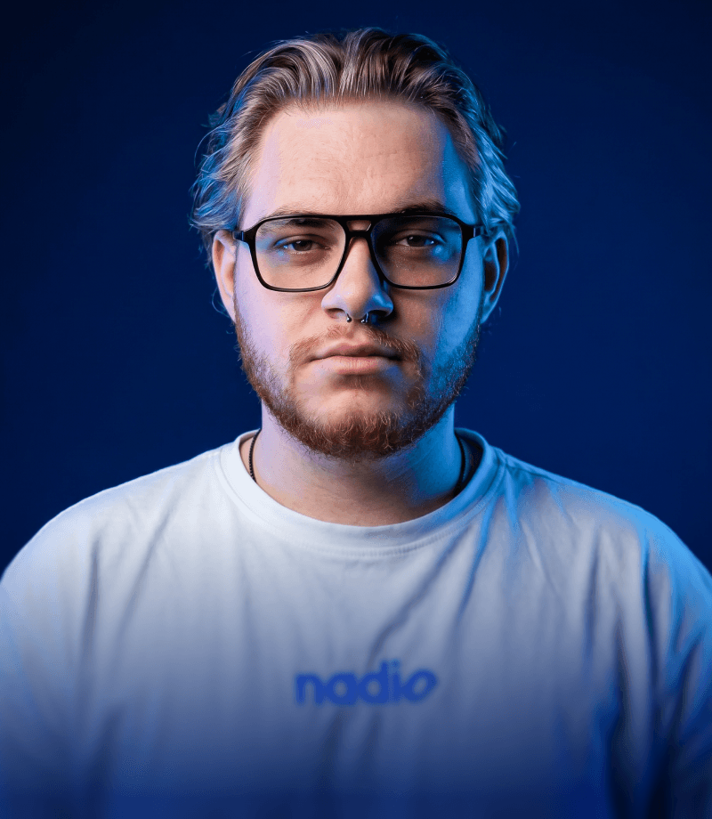 A young man with round glasses, a nose ring, and facial hair looks toward the camera against a dark, softly colored background.