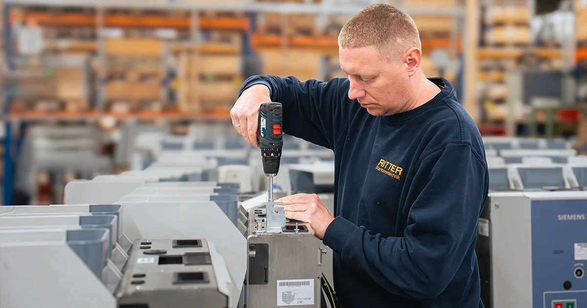 Ein Mitarbeiter von RITTER Starkstromtechnik montiert mit einem Akkuschrauber Komponenten an einem Elektroschaltschrank in der Fertigung. Im Hintergrund sind weitere Schaltschränke und der Produktionsbereich zu sehen.