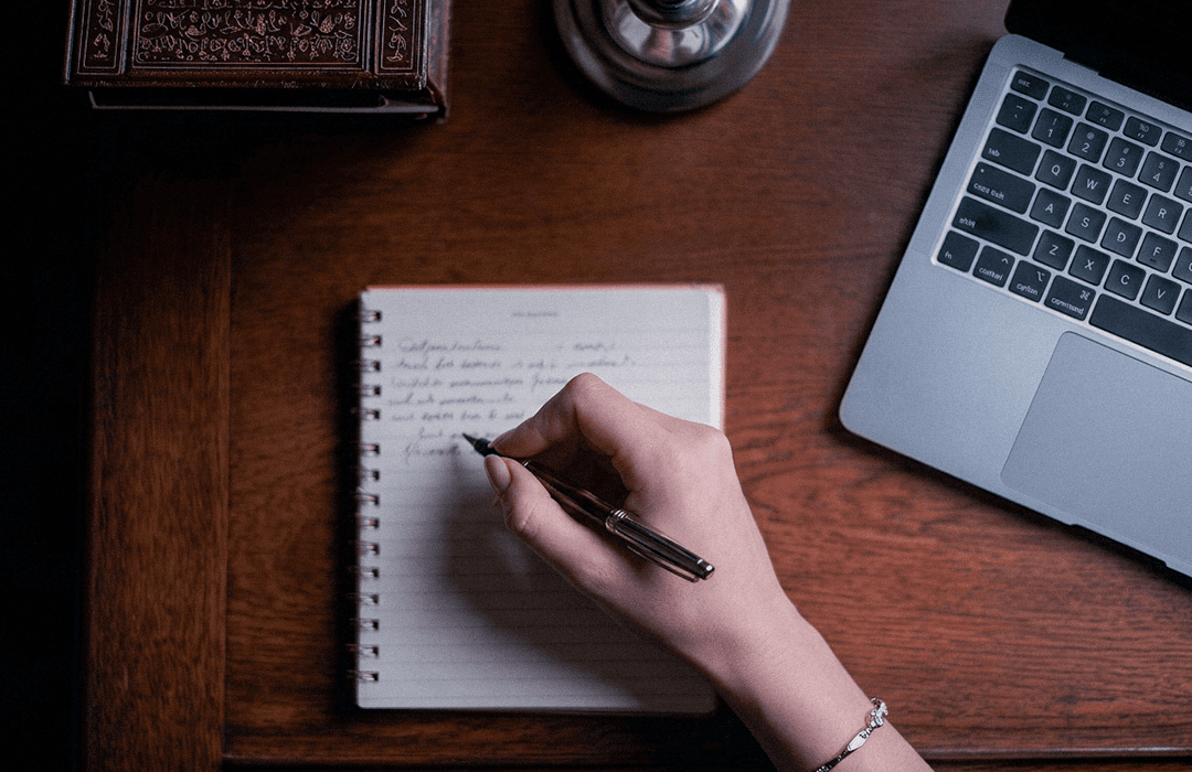 Vista superior de mão escrevendo em caderno ao lado de um notebook sobre mesa de madeira escura.