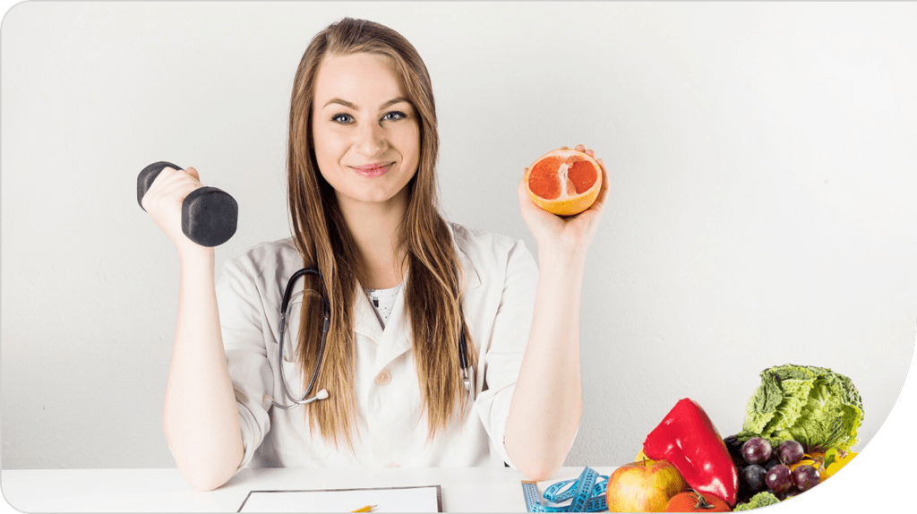 A woman in a white coat, with a stethoscope around her neck, holds a dumbbell in one hand and half a grapefruit in the other. On the table are fruits, vegetables, and a blue measuring tape, symbolizing her wellness-focused nutrition plan. She smiles at the camera.