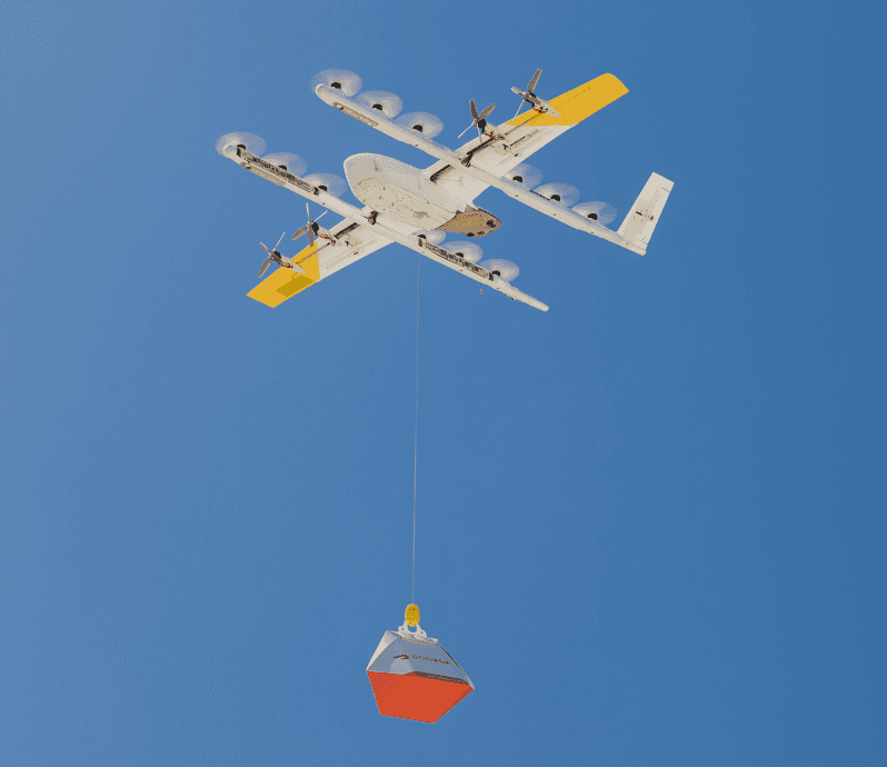 A white and yellow drone in flight against a clear blue sky; it is carrying a red and orange package, demonstrating modern delivery technology.