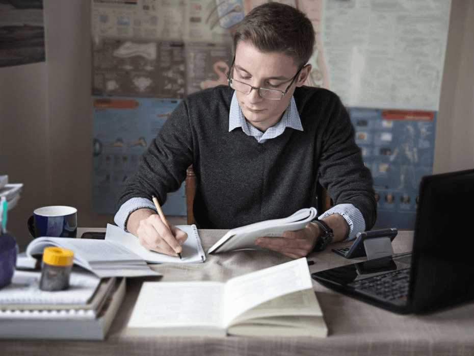 A high school student writing an abstract for a research paper at a desk with academic journals and a laptop