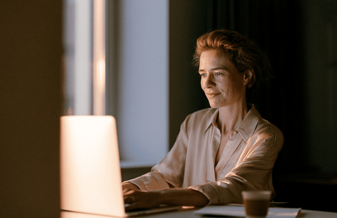 Mulher sorrindo levemente enquanto trabalha em um notebook à noite, com o rosto iluminado pela tela do aparelho. Blog post Inspira sobre agentes.
