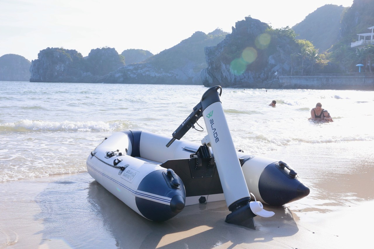 A Blade electric outboard motor tilted up out of the water on an inflatable boat beached on a sandy shore, with swimmers and limestone karst cliffs in the background.