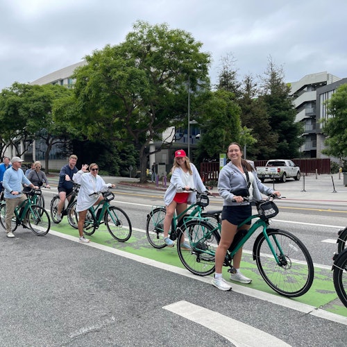 Group of people riding green rental bikes on a marked bike lane in an urban area with trees and buildings in the background.