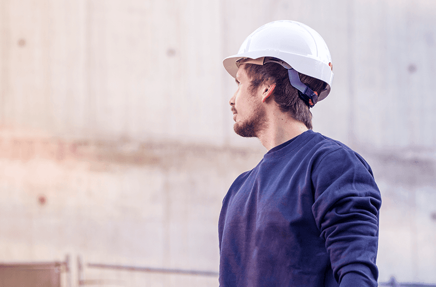 En arbeider iført hvit hjelm og marineblå genser ser til siden, stående foran en betongvegg