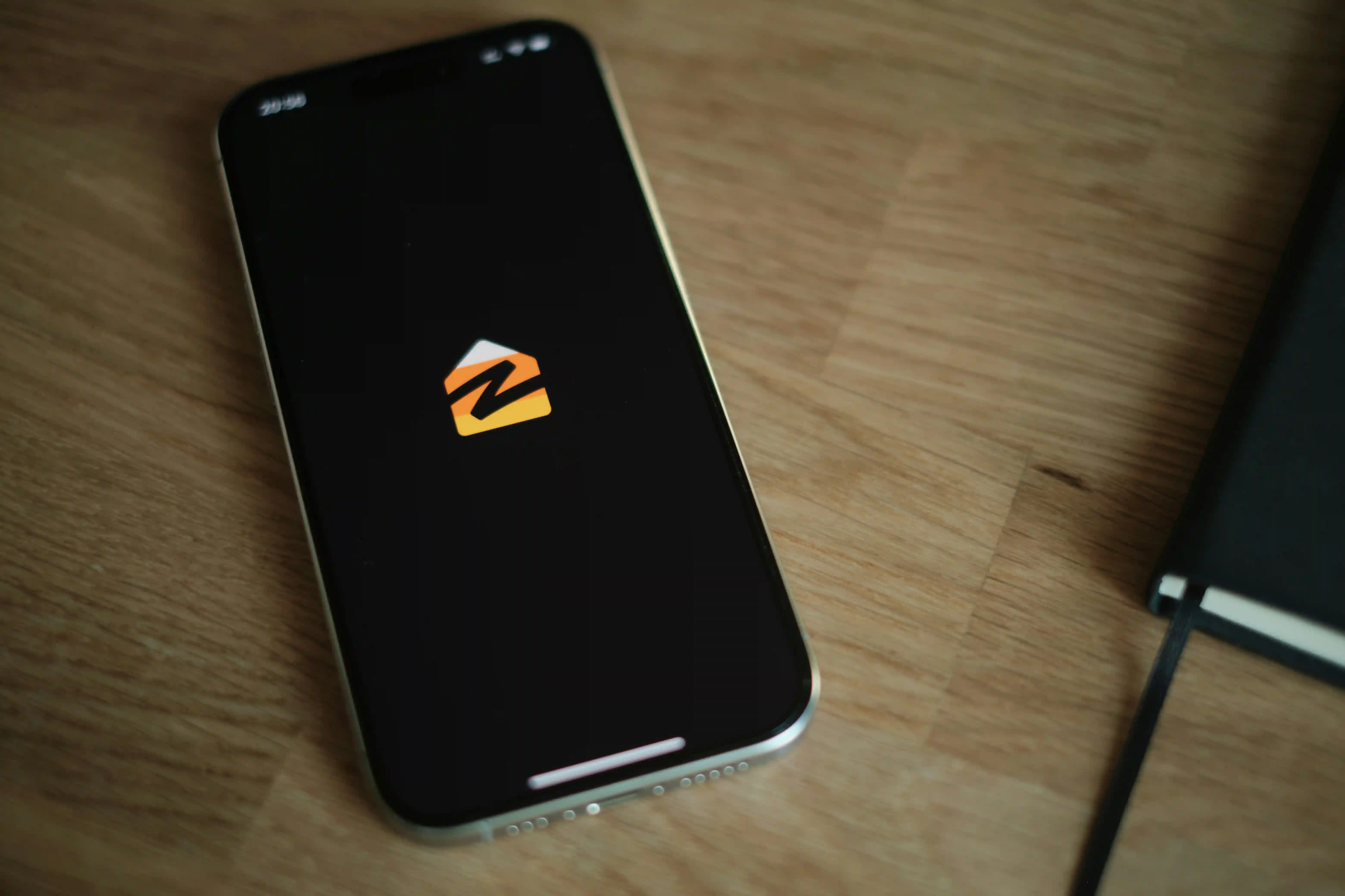 A smartphone displaying the Zillow app logo rests on a wooden desk next to a black notebook.
