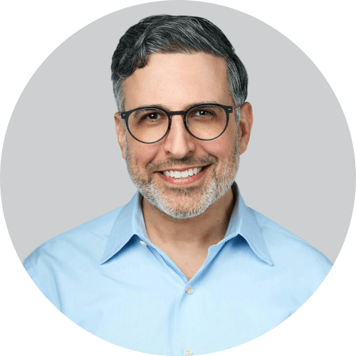 Portrait of a smiling man with glasses and short gray hair, wearing a light blue shirt.