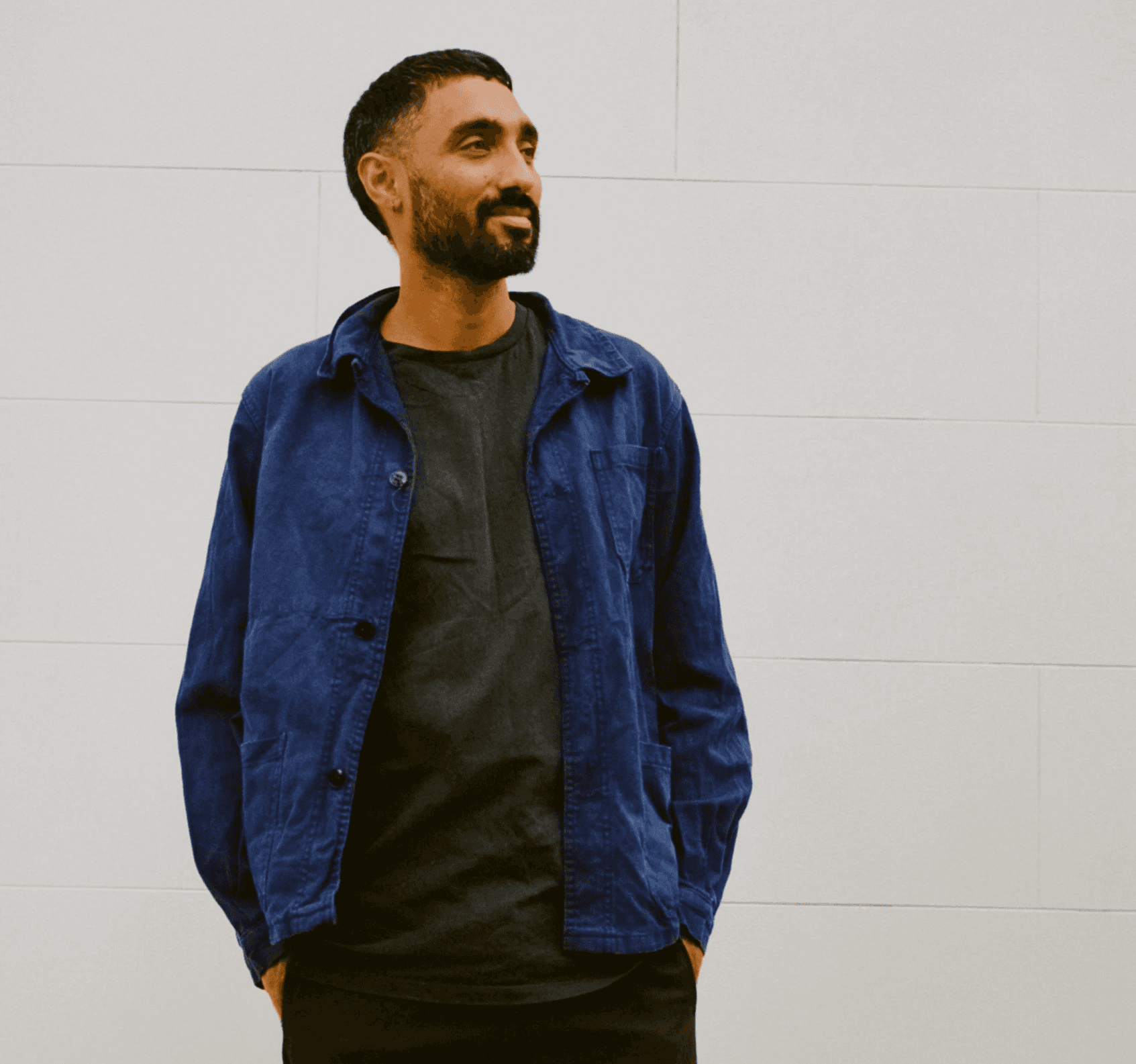 Man with dark hair and beard wearing blue jacket standing in front of a white wall