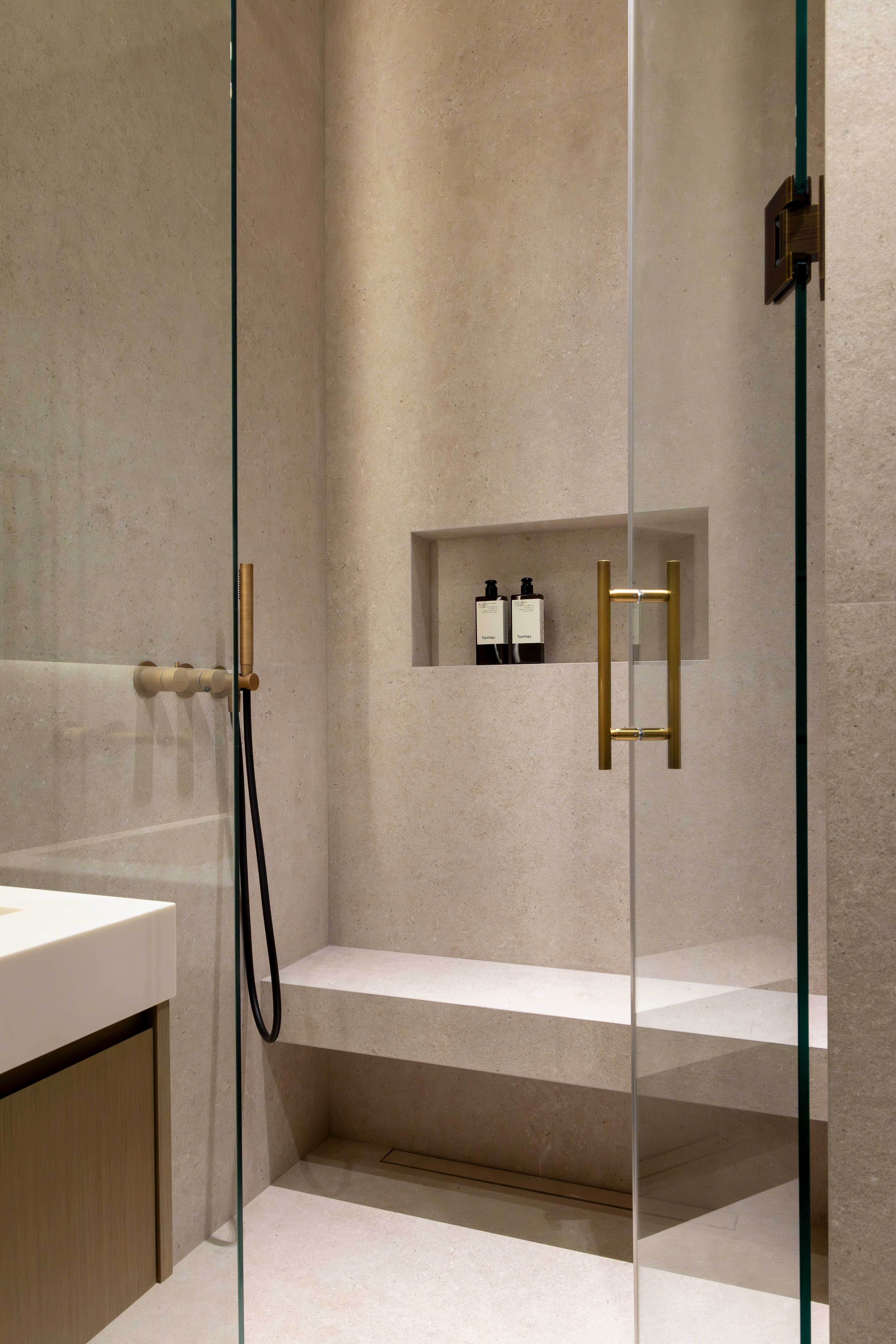 Douche italienne en pierre beige avec banc integre, niche de rangement, robinetterie doree et paroi en verre
