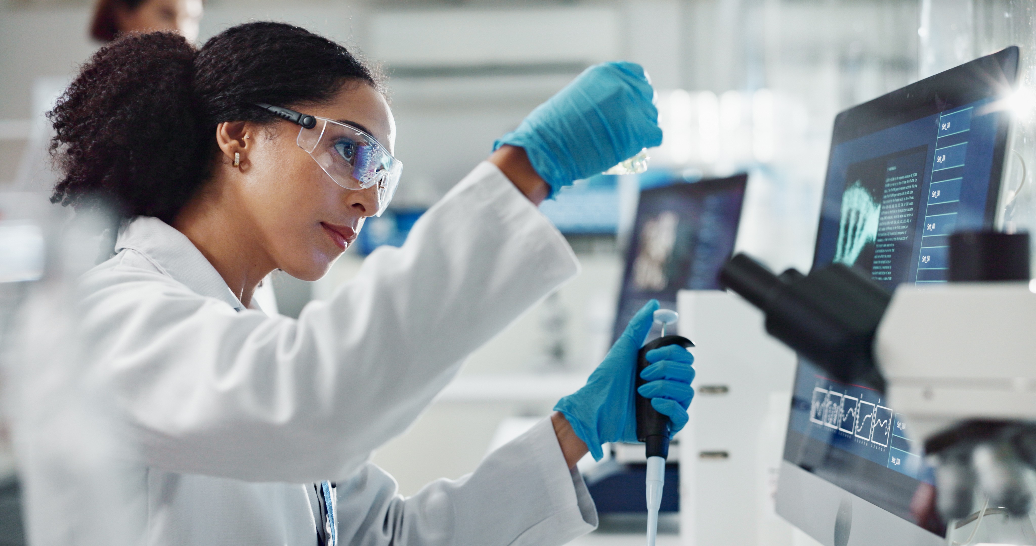 A female scientist wearing lab goggles and gloves holding a vial and pipette.