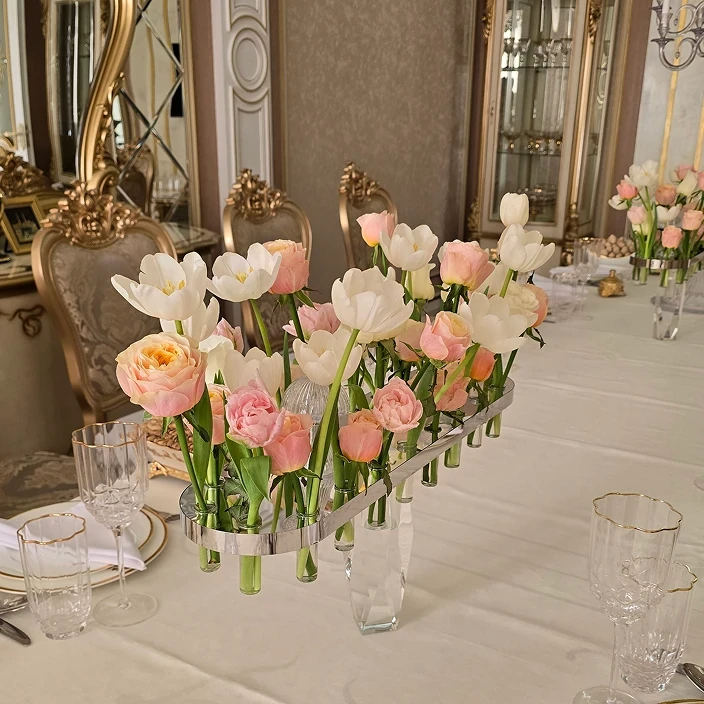 Elegant dining table with white tulips and pink roses in a modern glass centerpiece, surrounded by gold-detailed chairs in a luxurious setting.