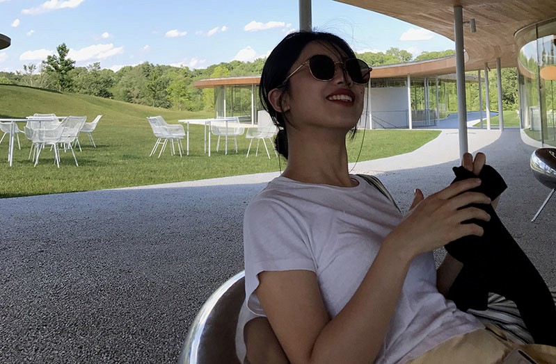 A photo of a girl wearing a white t-shirt and sunglasses, sitting in an open terrace. In the background there is a forest, some green grass, some white tables and chairs.