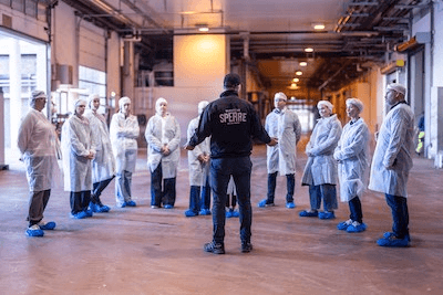 A group of workers in protective gear listens attentively to a speaker in a large industrial setting.