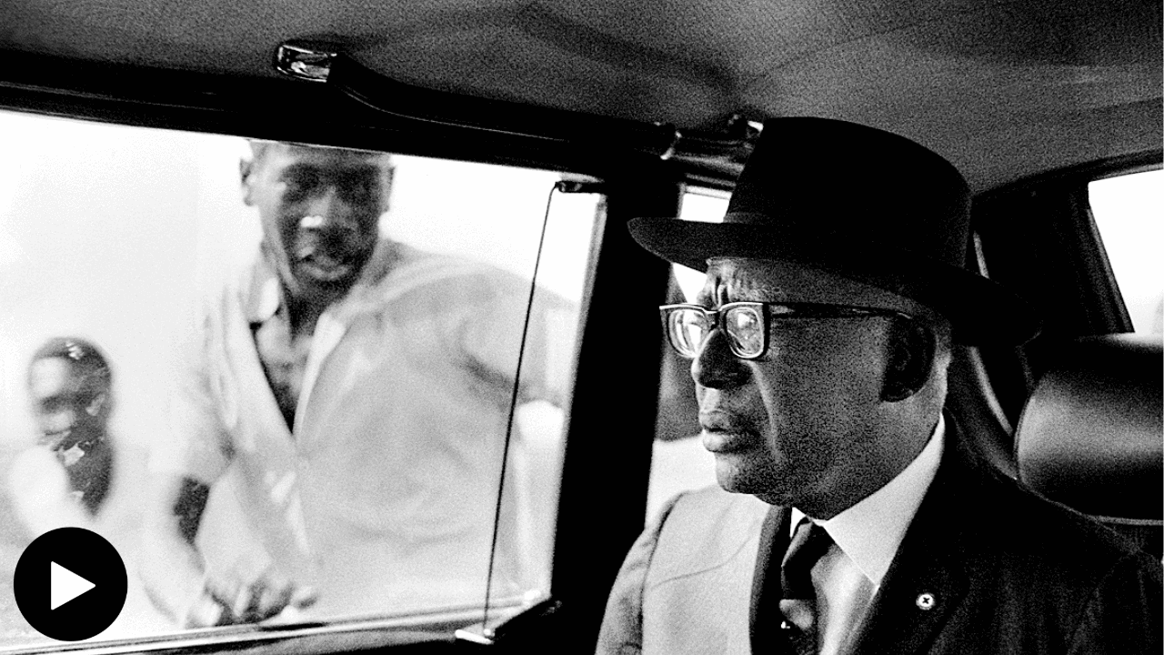 A black and white photograph of François Duvalier, the president of Haiti from 1957 until his death in 1971. He looks out the window of the car he's seated in, two men running alongside the automobile. They are dressed in white button-up shirts, contrasting Duvalier's black suit and hat.
