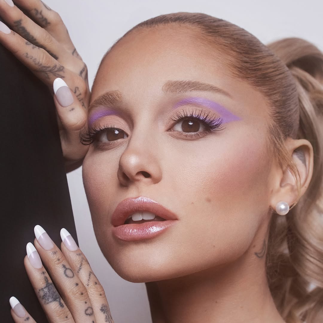 Ariana Grande wearing lavender eyeshadow and glossy lips, posing with pearl earrings and manicured nails for r.e.m. beauty.
