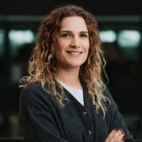 A smiling person with long, curly hair stands with arms crossed, in a modern, blurred background.