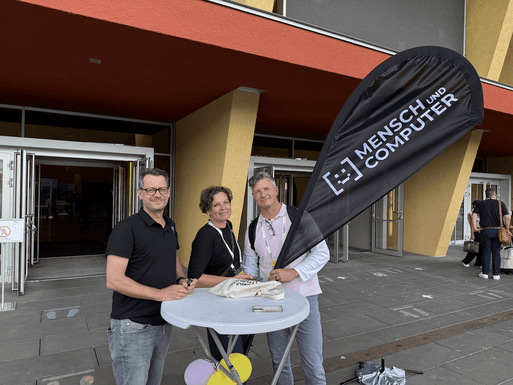 Drei Personen stehen an einem Stehtisch vor einem Gebäude, daneben ein schwarzer Fahnenbanner mit der Aufschrift 'MENSCH UND COMPUTER'