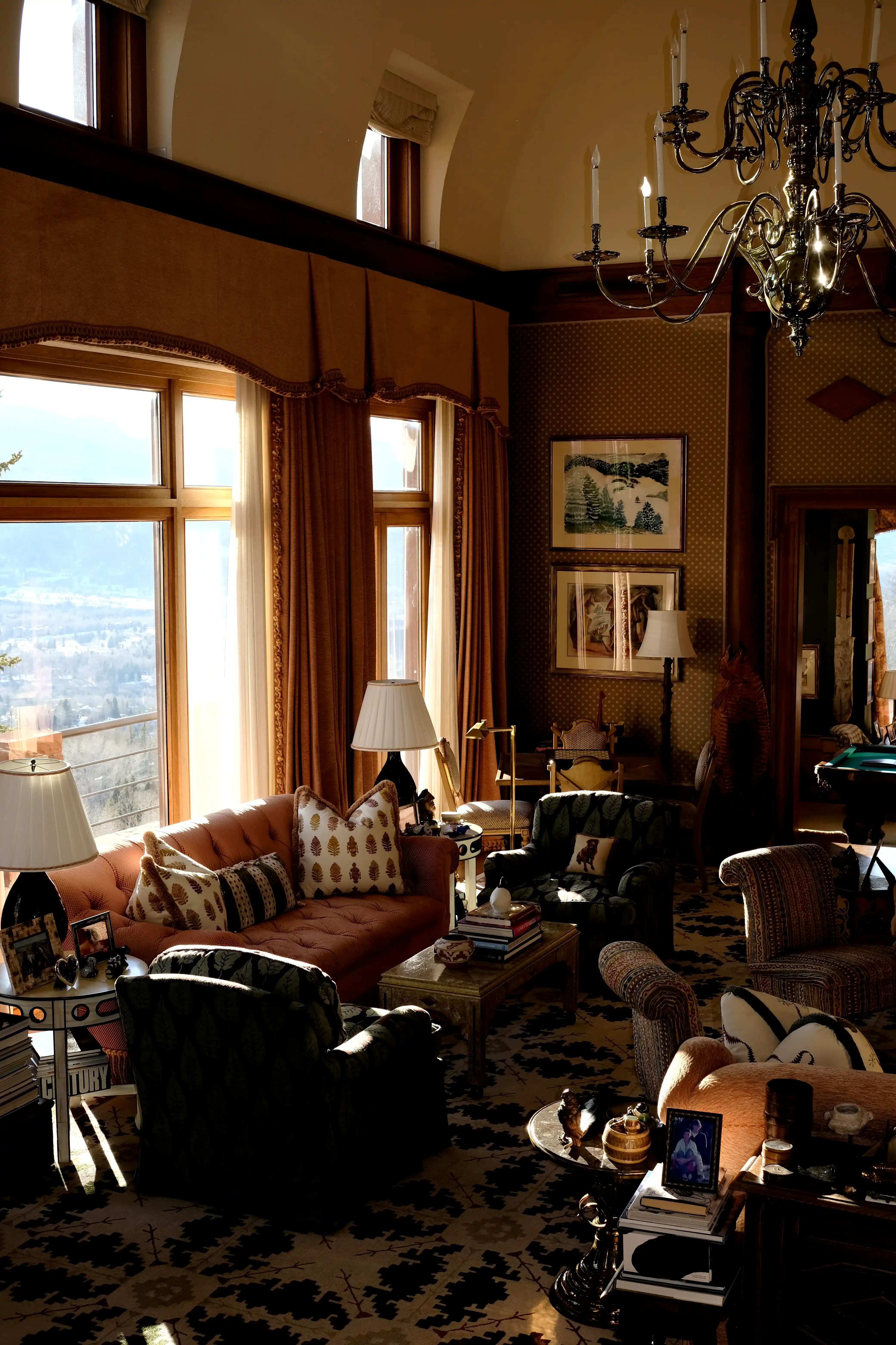 Sunlit luxury mountain lodge living room with tall windows, ornate chandelier, patterned rugs, sofas, armchairs, and warm wood interiors.