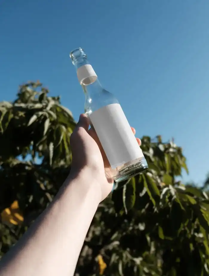 Clear glass bottle mockup held up against a bright blue sky with tree foliage in the background. The minimalist label design emphasizes a refreshing, natural aesthetic in an outdoor setting.