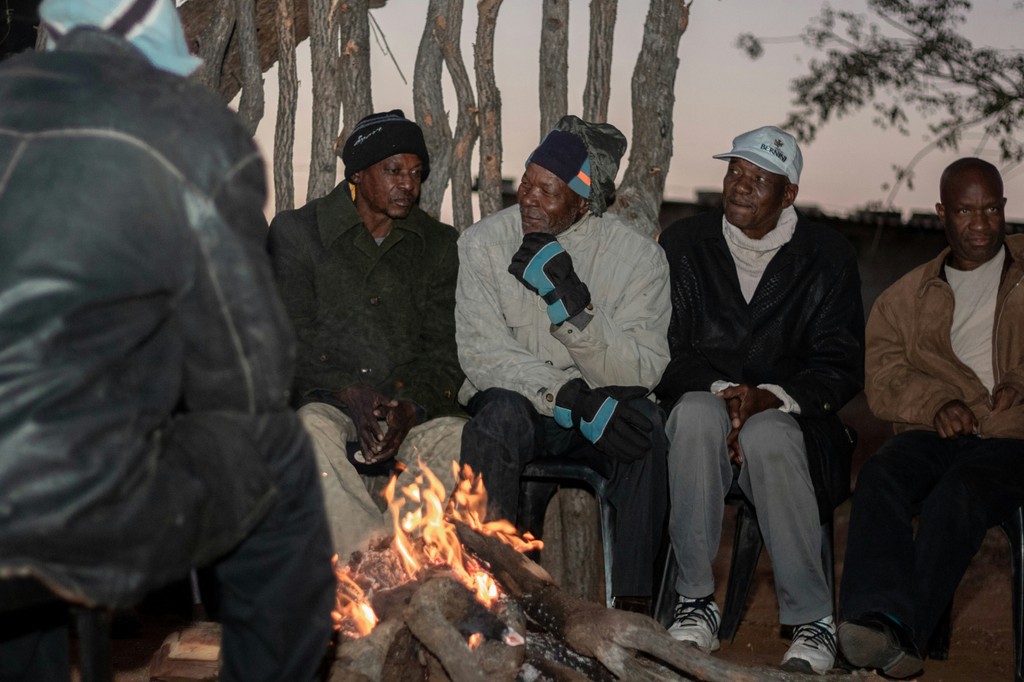 A group of elderly men wearing winter clothing sit around a warm, crackling campfire, engaged in conversation against a backdrop of wooden branches and a dusky sky.