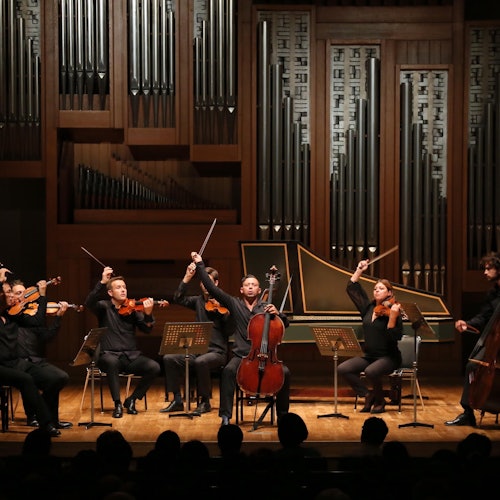 A group of orchestral musicians on stage, playing string instruments in front of a large pipe organ. Audience visible in the foreground.