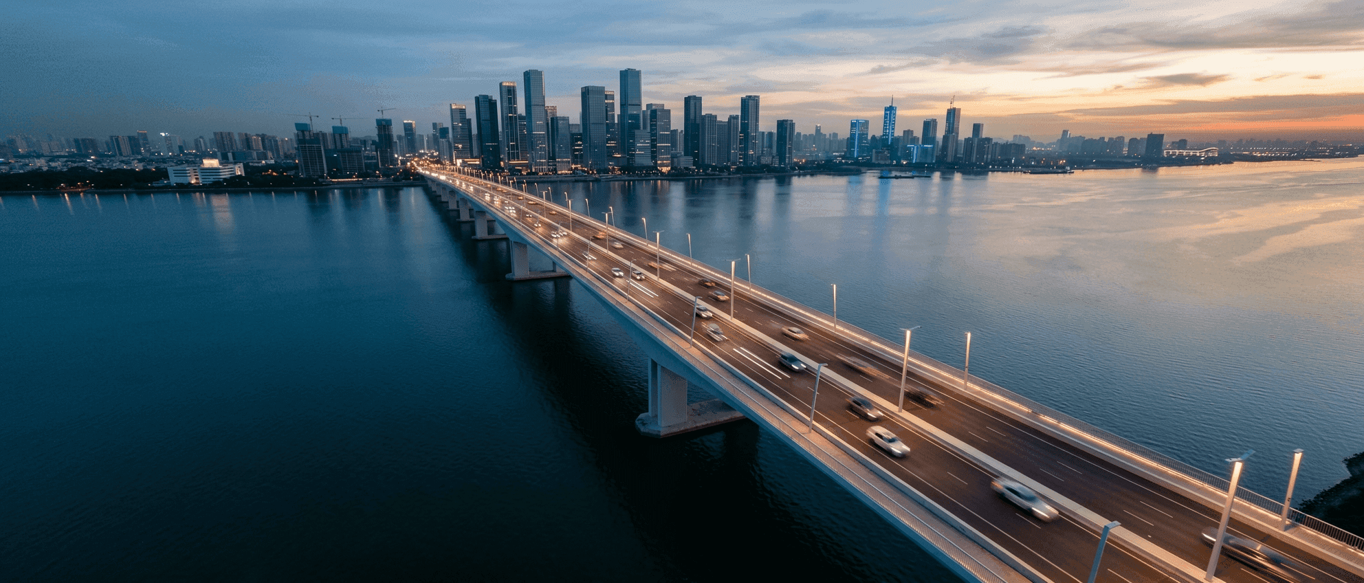 Aerial view of a highway bridge over a lake at dusk, city skyline in the background