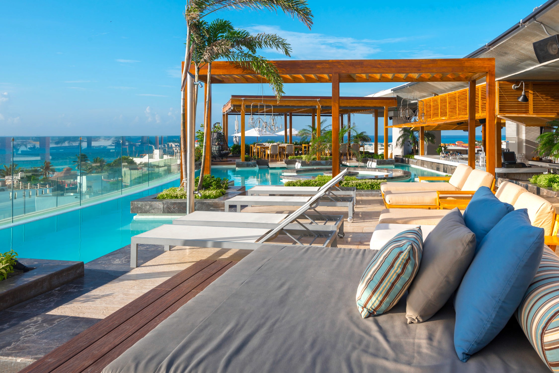 A luxury poolside lounge area with white and blue cushions features a wooden cabana and an infinity pool overlooking the