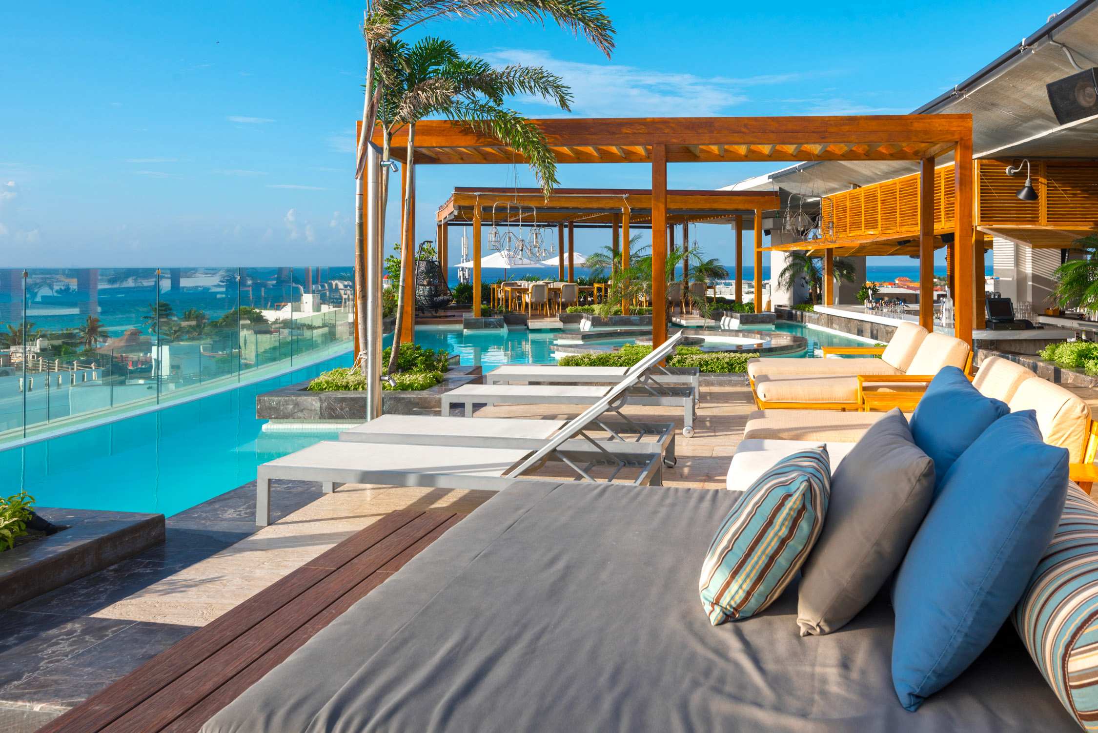 A sunlit poolside lounge features a wooden pergola with white cushions and blue pillows overlooking a turquoise pool and ocean.