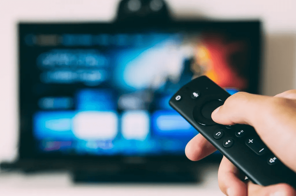 A hand holding a remote control, which is controlling a streaming service on a distant TV.