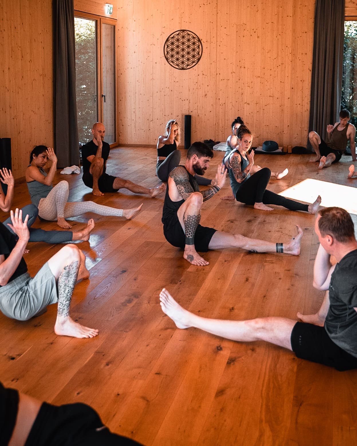 Flo instructing a group of yoga teacher students on the mat in a wooden sunlit studio.
