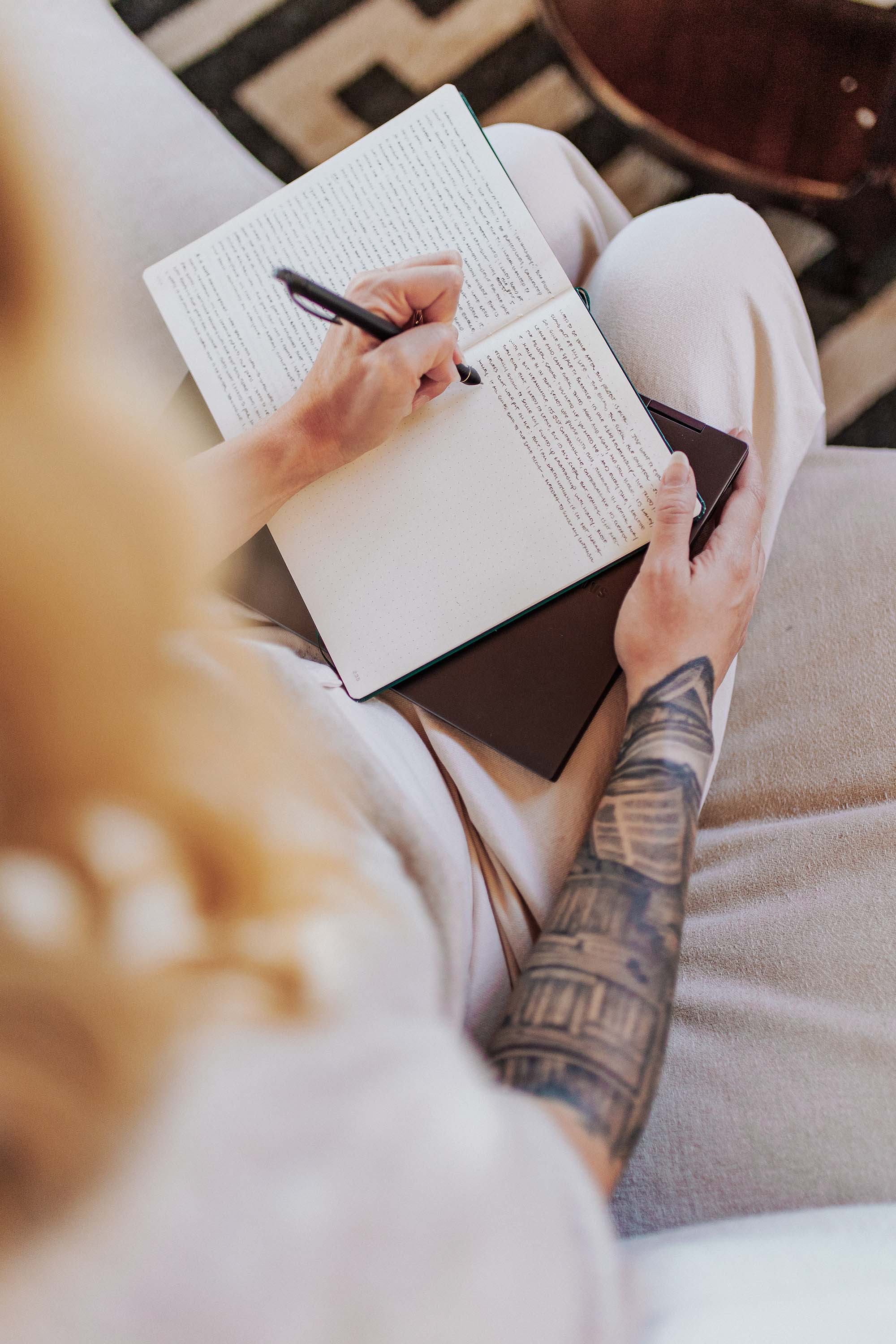 A top down view over the shoulder of a woman sitting on a couch and writing in a journal