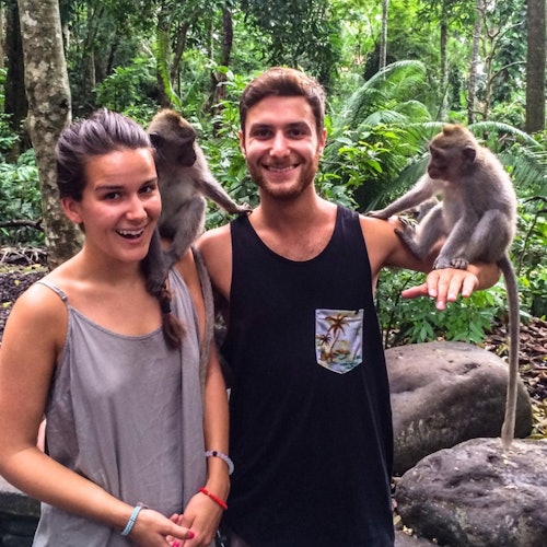 Two people smiling with monkeys on their shoulders and arm in a lush, tropical forest setting.