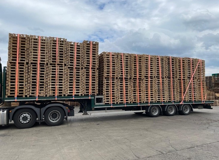 A truck with a flatbed trailer is loaded with neatly stacked wooden pallets, secured with orange straps, on a concrete surface under a cloudy sky.