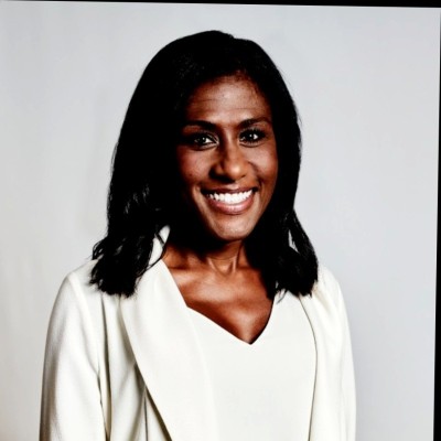 Smiling woman wearing glasses and a pink blazer on a warm pastel background.
