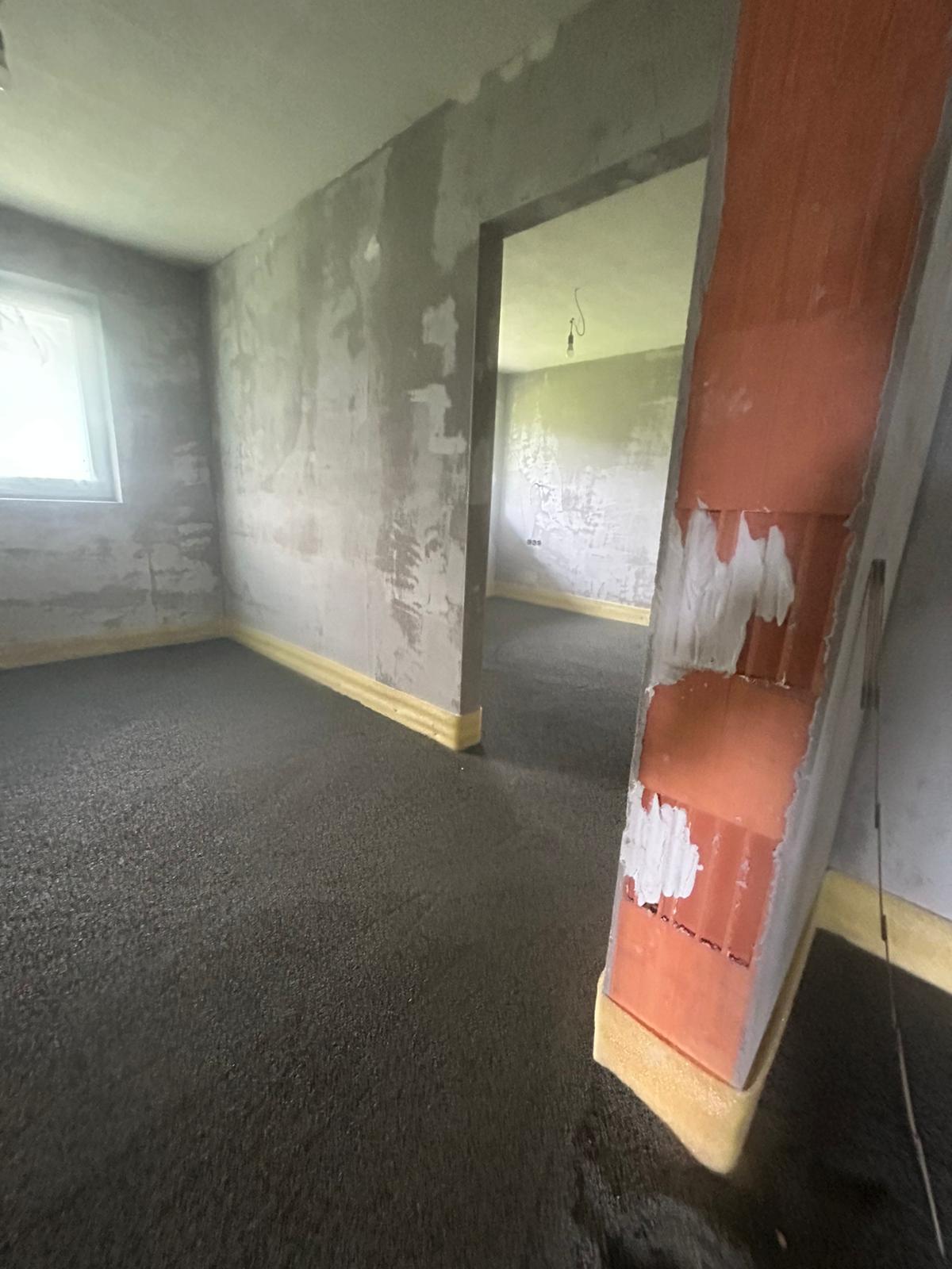 Innenansicht eines im Bau befindlichen Raumes mit Betonboden, Türöffnung und freiliegender Ziegelsäule.
