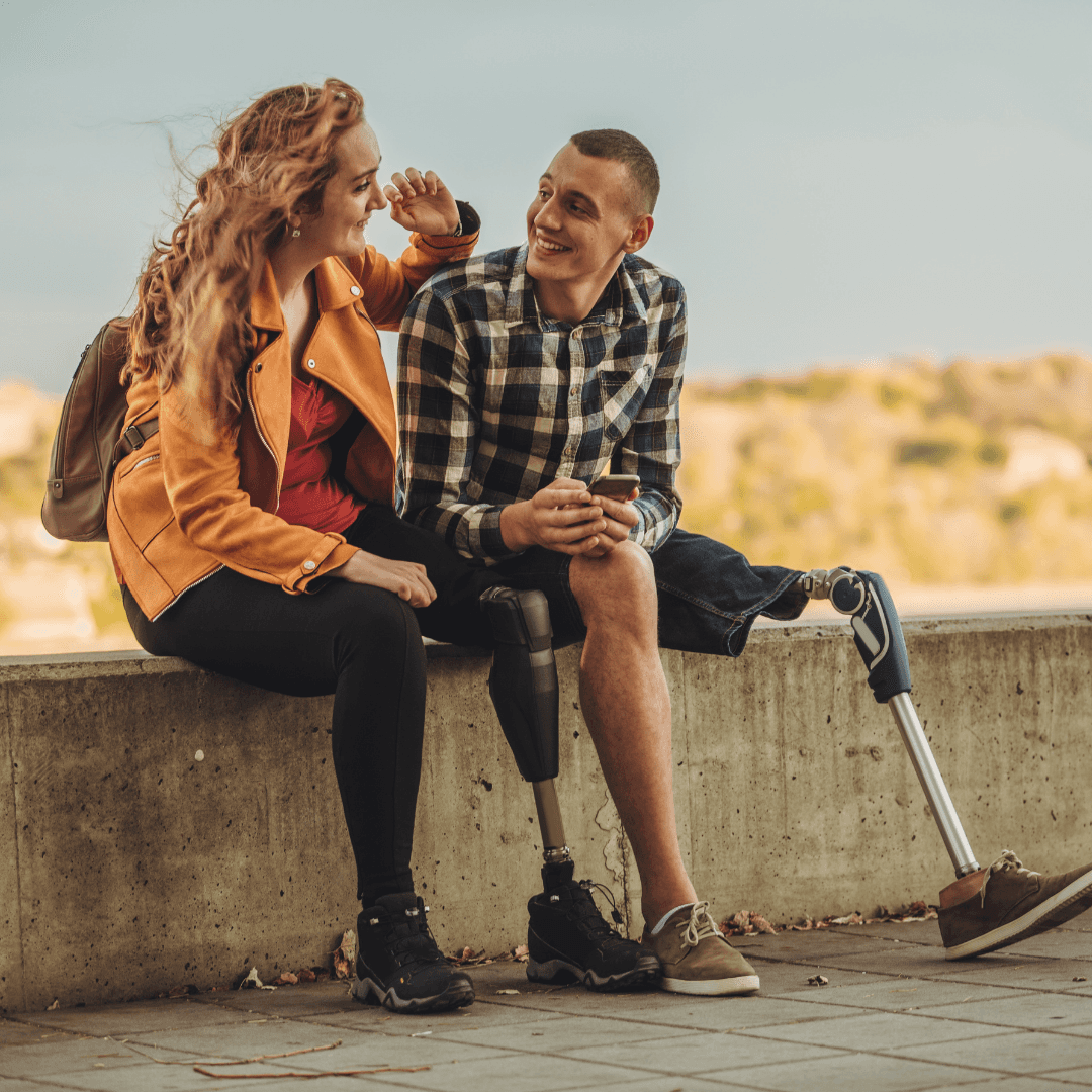 A woman and man with prostetic legs sitting on a ledge holding a smartphone