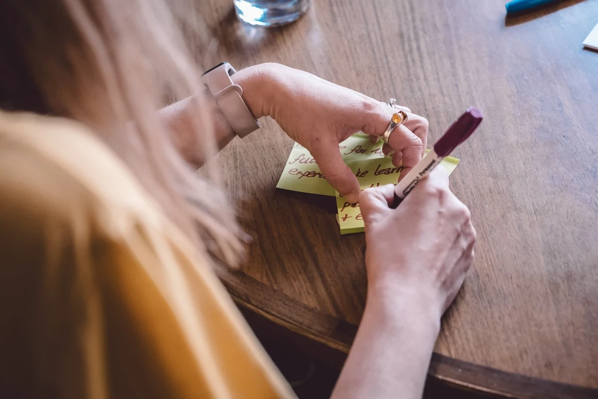 Close-up of hands writing on a yellow sticky note with a marker at a wooden table, with another note partially visible underneath.
