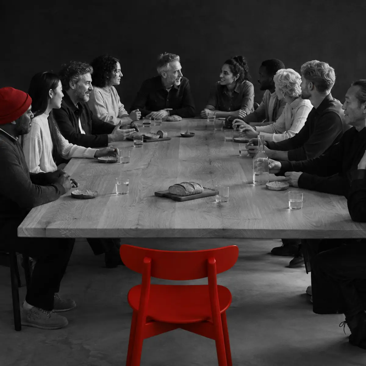 People of different ages and backgrounds gather around a table with one open red chair.