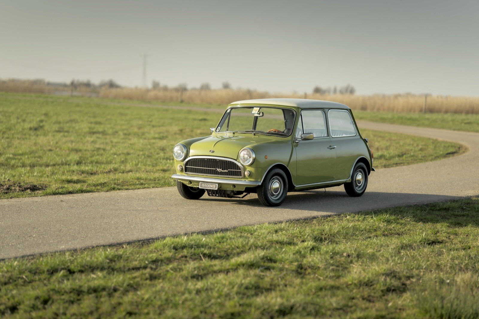 A vintage green car parked on a winding road with open fields in the background under a clear sky.