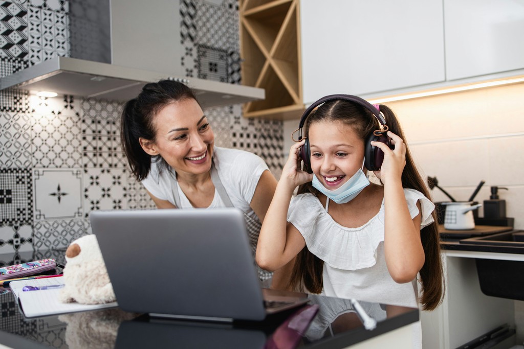 A smiling mother watches her daughter, who is wearing headphones and a face mask, as she uses a laptop in a modern kitchen, highlighting the blend of remote learning and parental support during the pandemic.